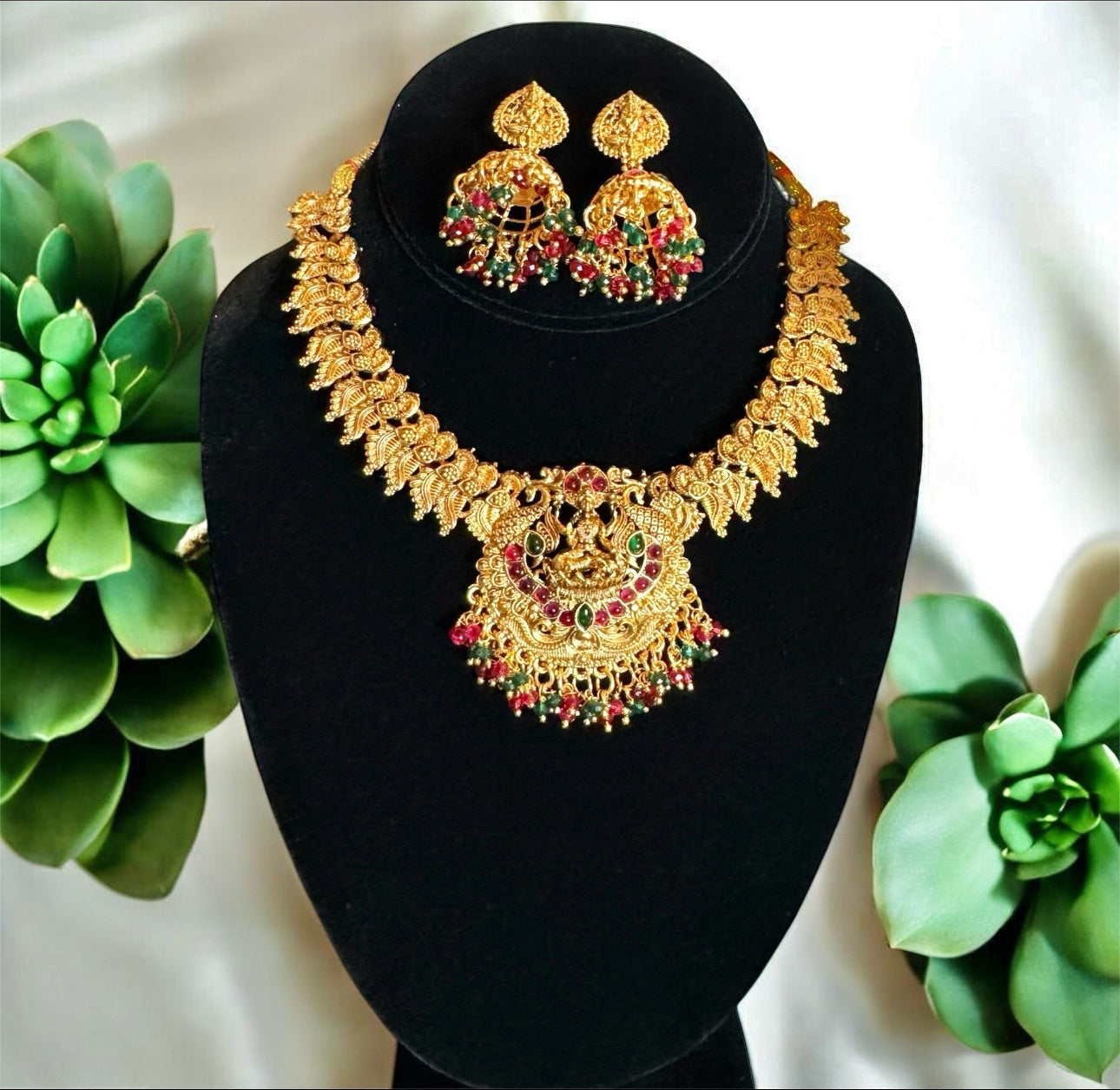 Gold necklace and earrings set on a black stand with green plants in the background