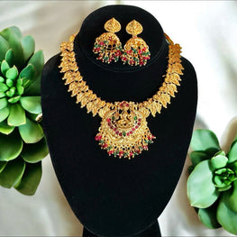 Gold necklace and earrings set on a black stand with green plants in the background